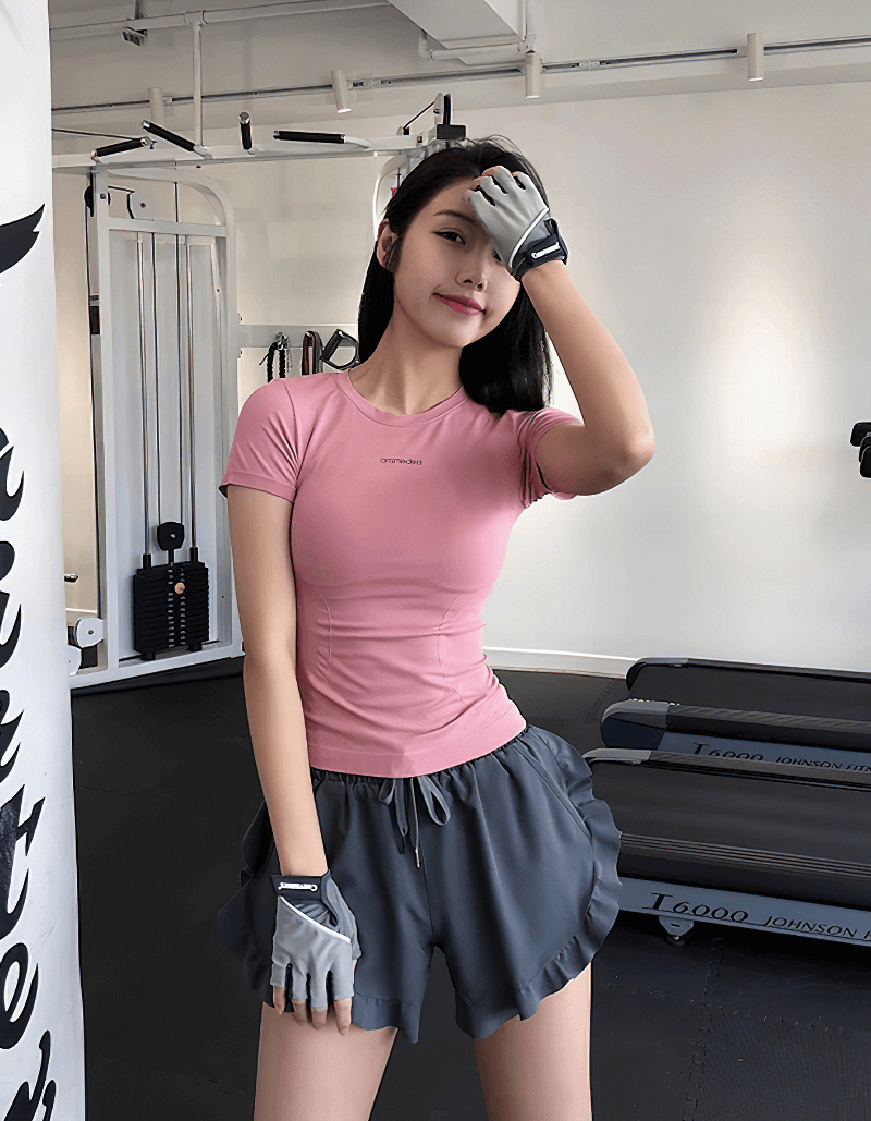 Woman wearing a pink breathable short sleeves fitness t-shirt and gray shorts in a gym, showcasing active sportswear style.